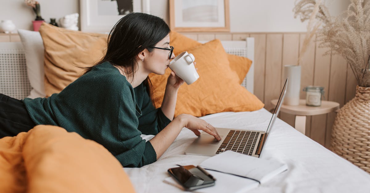 Woman relaxing on bed with coffee browsing laptop for freelance work or online learning.